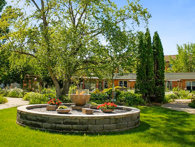 Outdoor courtyard with circular stone fountain, trees, and flowering plants in front of a brick building.