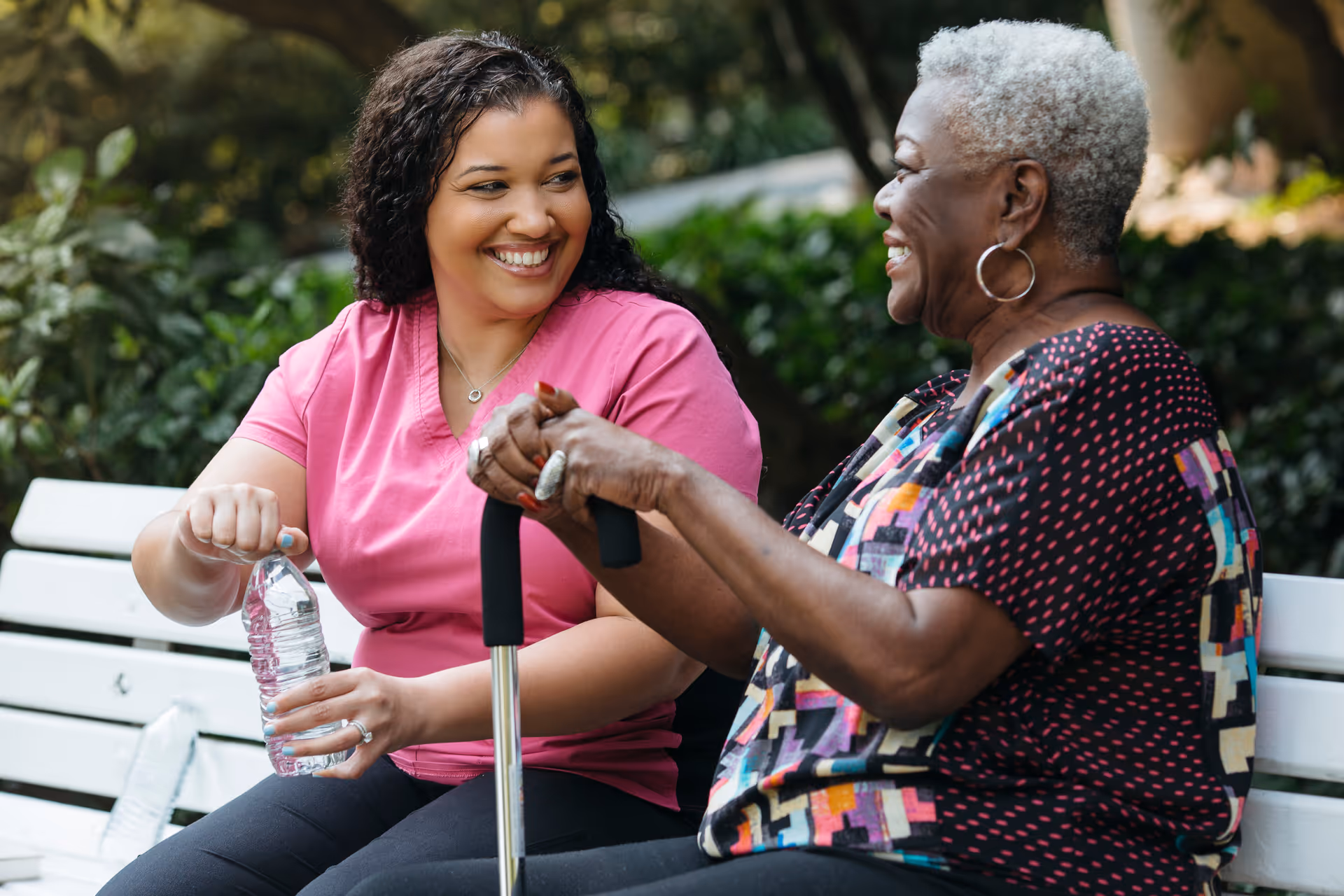 Caregiver and senior woman smile while seated on an outdoor bench, sharing a friendly moment.