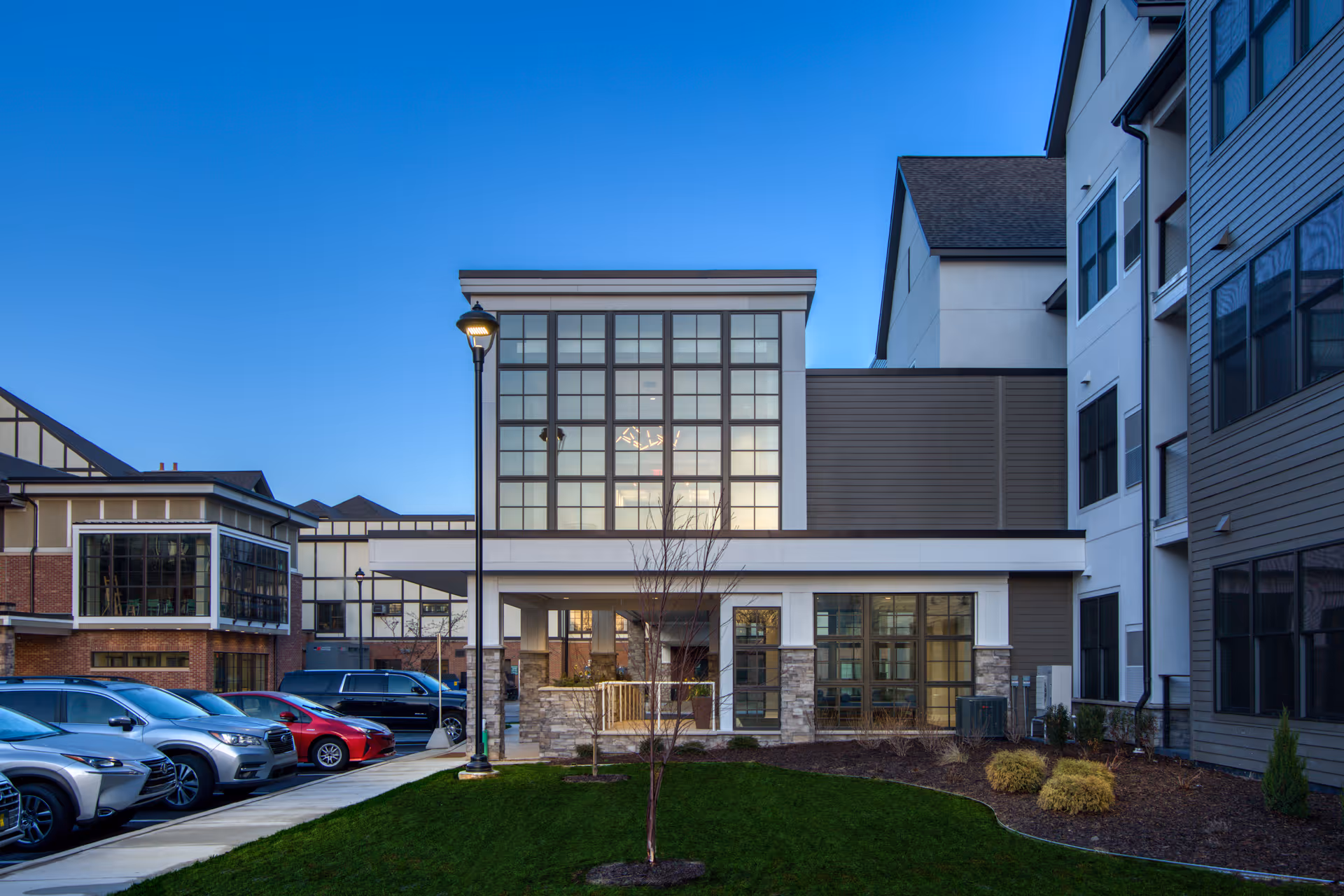 Exterior view of a modern senior living facility building with large windows, a well-maintained lawn, and a parking area with several cars parked. The sky is clear and blue.