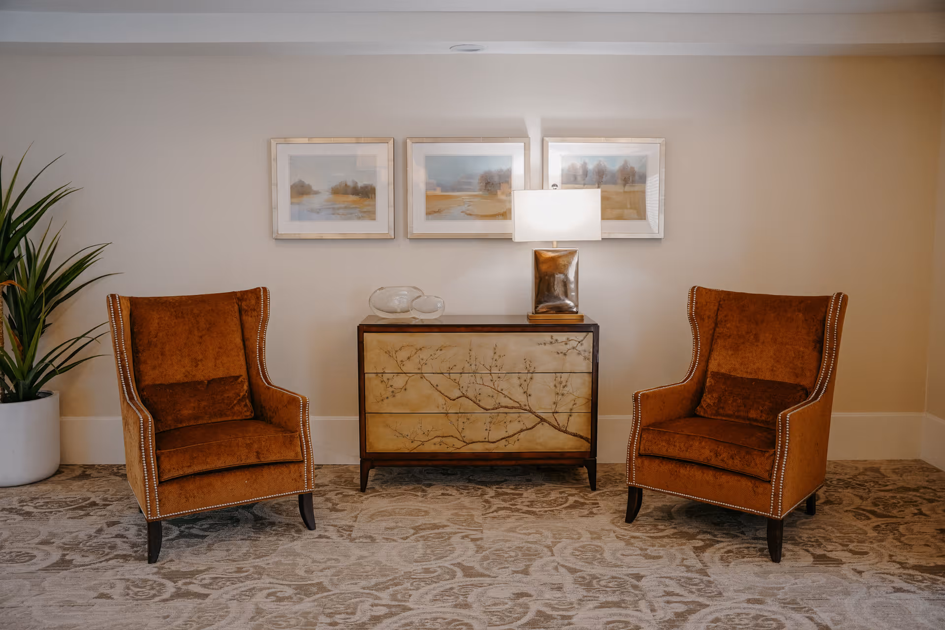 Two brown upholstered armchairs with nailhead trim facing forward, positioned on either side of a wooden chest of drawers with a floral branch design. On top of the chest is a modern table lamp with a rectangular base and a white shade, along with two decorative glass objects. Above the chest are three framed landscape paintings. A large potted plant is visible to the left, and the floor is covered with a patterned carpet.