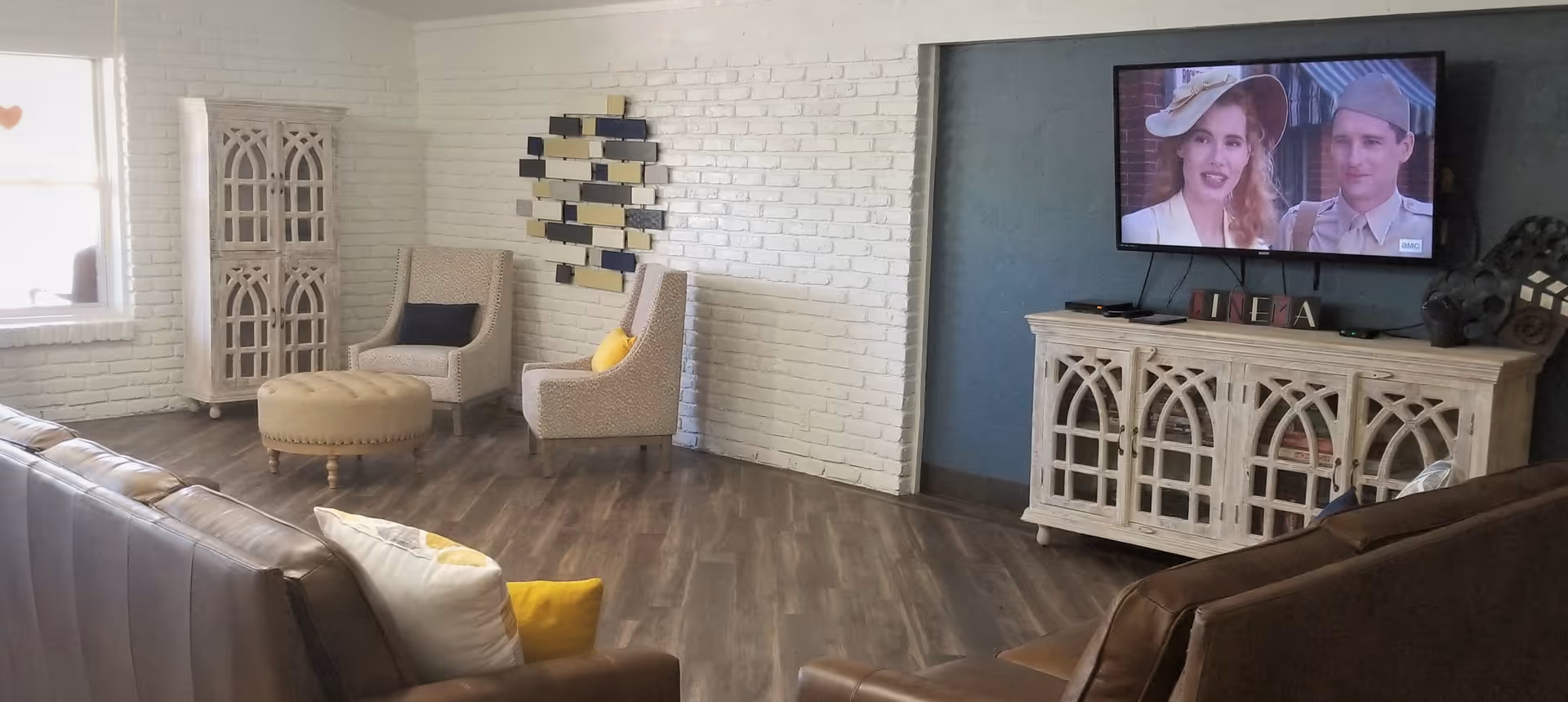 A cozy living room with brown leather sofas, two beige armchairs with yellow and dark blue cushions, a round beige ottoman, a white brick wall, a decorative wall art piece, a white wooden cabinet with glass doors, and a flat-screen TV mounted on a blue wall showing a scene from a movie or TV show.