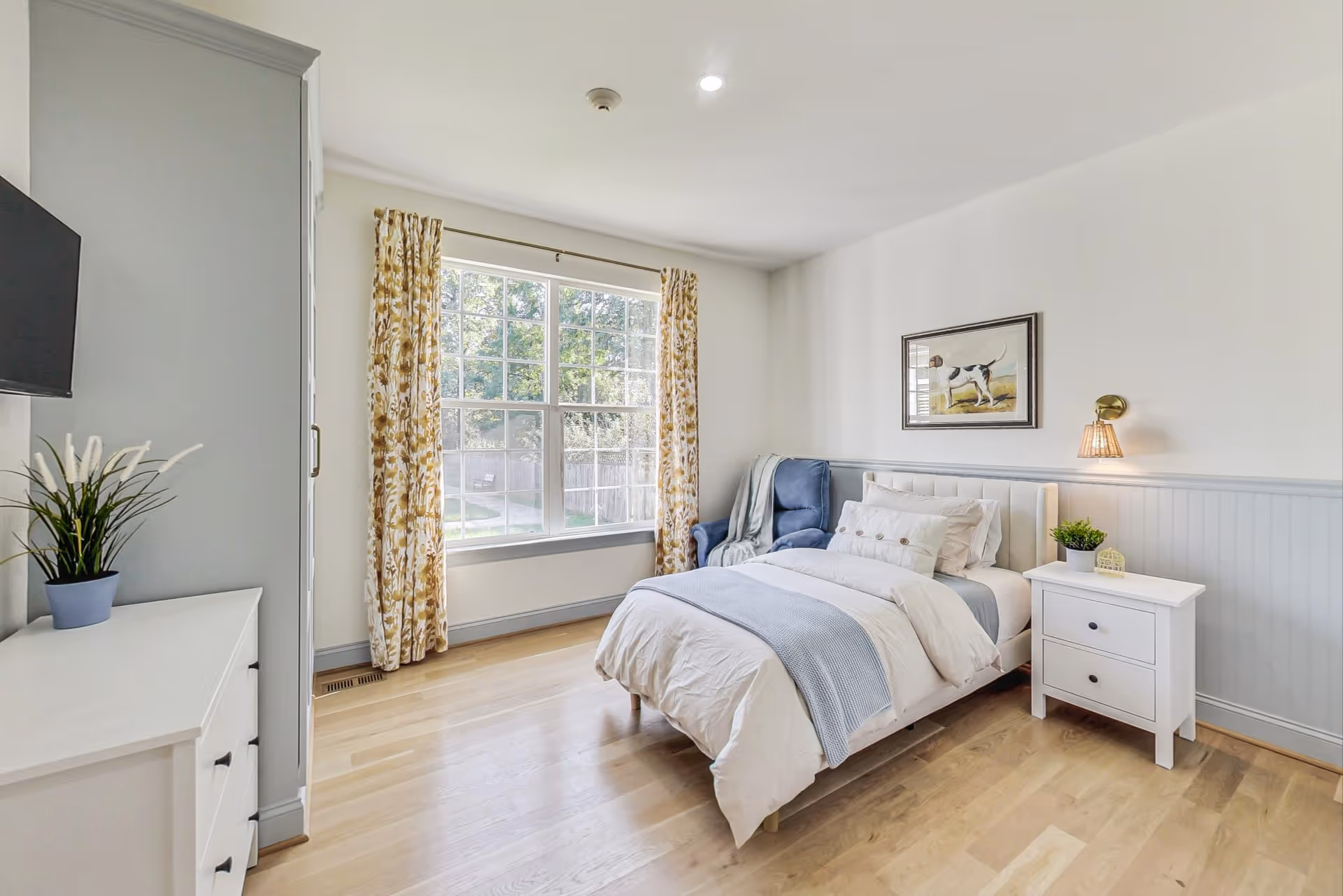 Bright bedroom in a senior living facility with a bed, dresser, window, and floral curtains.