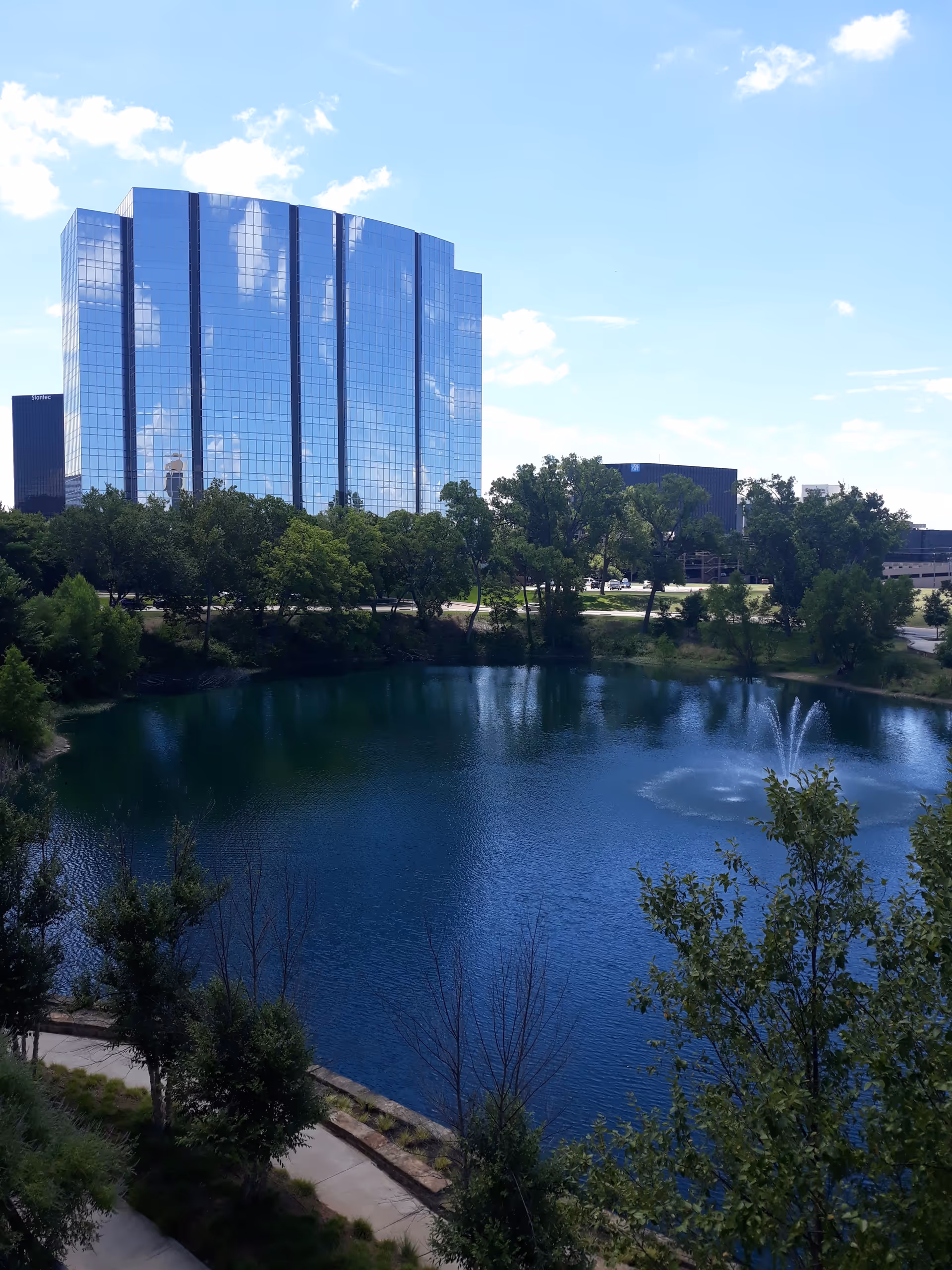 A tall glass building stands beside a tree-lined pond with a fountain.