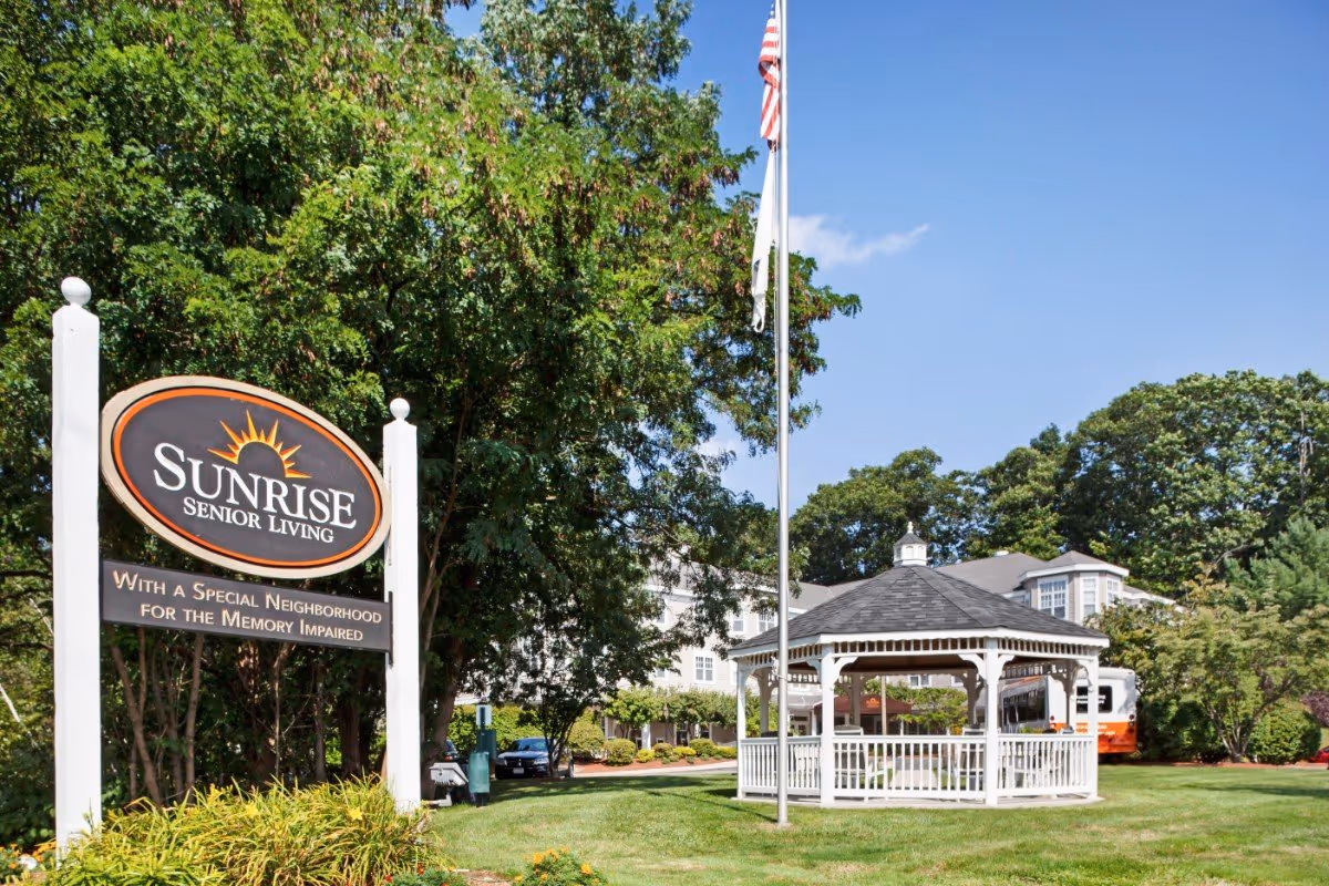 Entrance sign for Sunrise Senior Living with a gazebo, flagpole, and the facility building in the background.