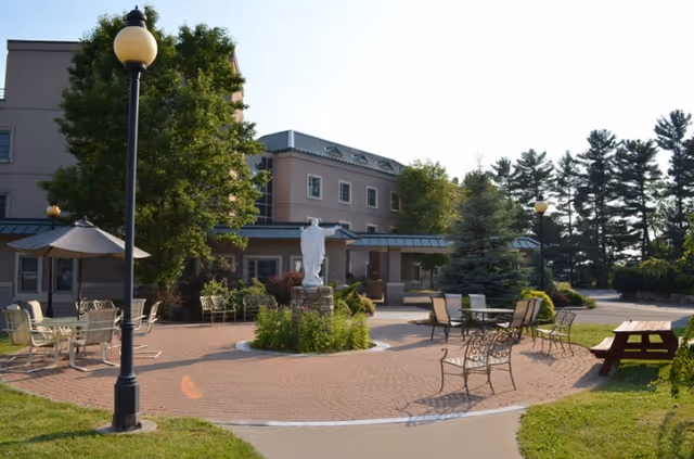 Outdoor patio area at Capstone Center for Rehabilitation and Nursing featuring a circular brick-paved seating area with metal chairs and tables, a statue in the center surrounded by greenery, lamp posts, trees, and a multi-story building in the background under a clear sky.