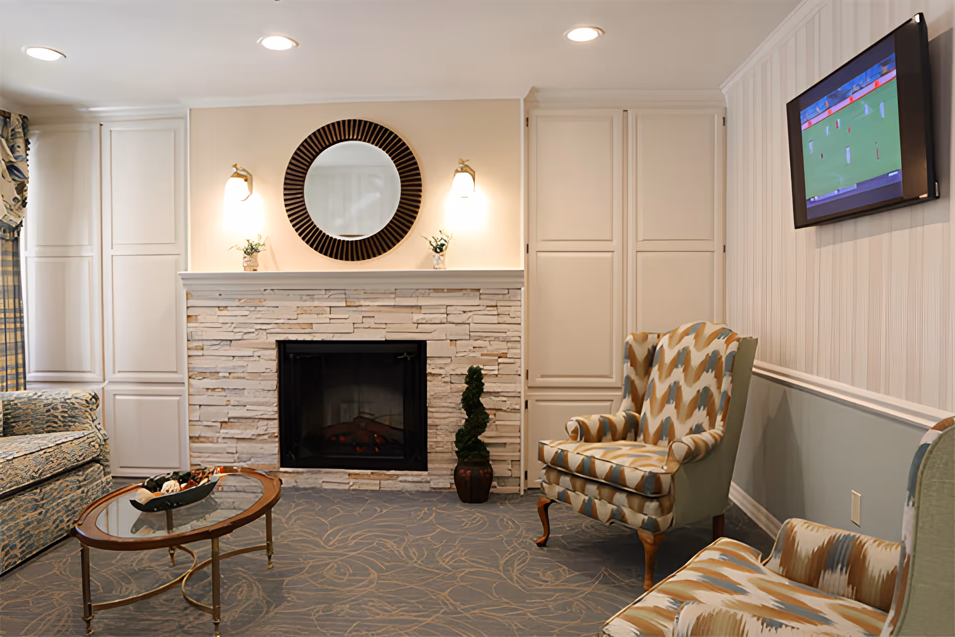 Comfortable living room with a stone fireplace, round mirror, patterned armchairs, glass coffee table and wall-mounted TV.