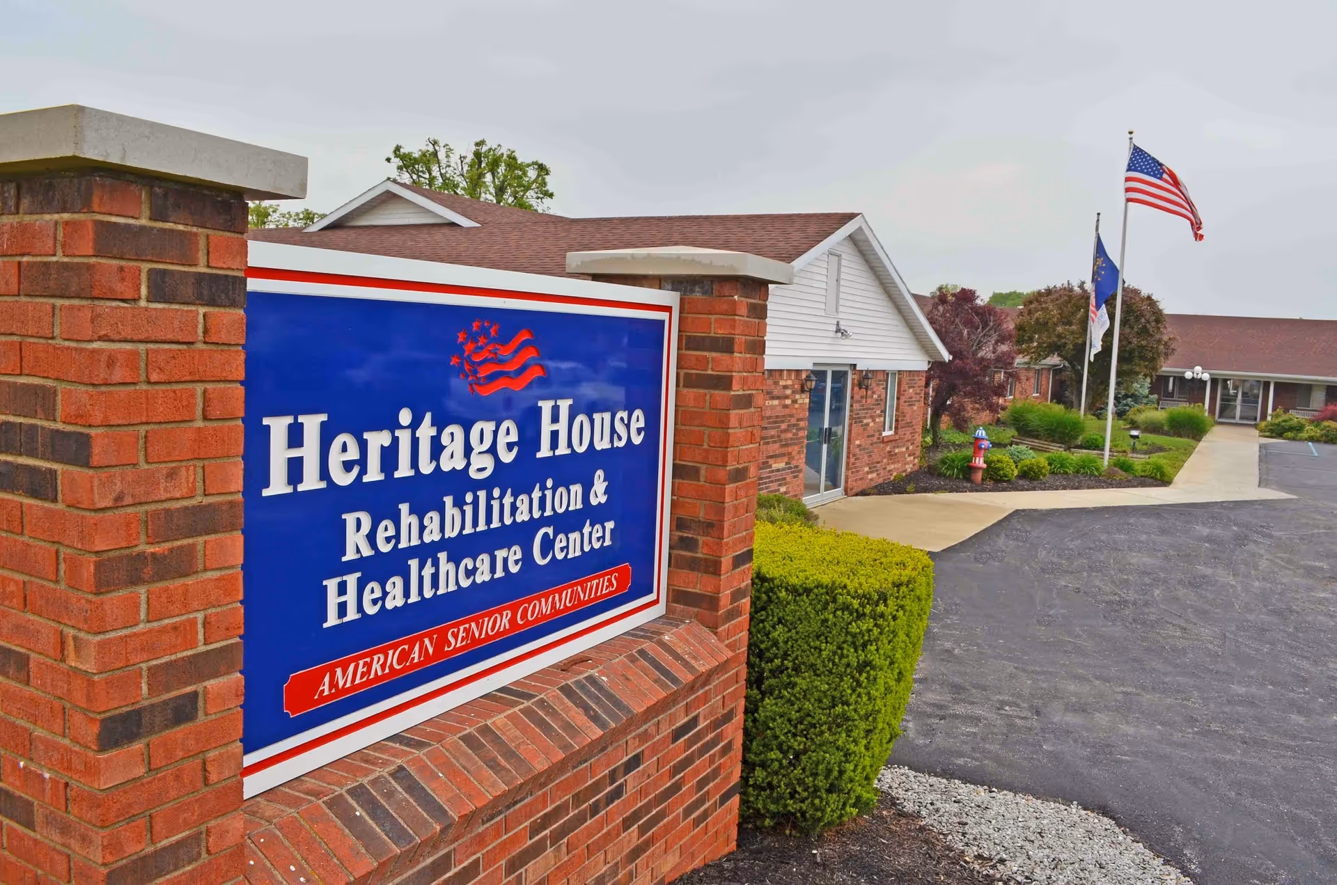 A brick sign reading Heritage House Rehabilitation & Healthcare Center outside the facility with the building entrance, landscaping, and American flags in view.