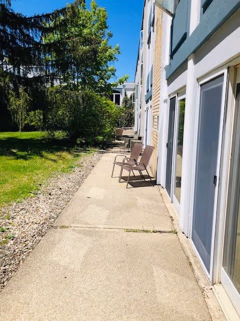 Outdoor walkway alongside a building with several chairs placed against the wall. The building has multiple doors and windows, and there is a grassy area with trees on the left side under a clear blue sky.