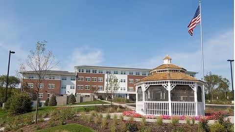 Front exterior of a multi-story senior living building with a white gazebo, landscaped grounds, and an American flag.