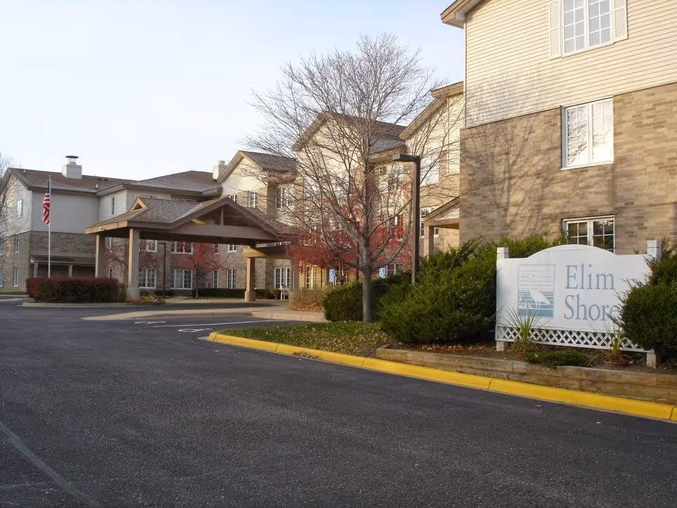 Exterior view of a senior living facility named Elim Shores, showing a multi-story building with beige siding and brick accents, a covered entrance, leafless trees, bushes, and a paved driveway with a yellow curb.