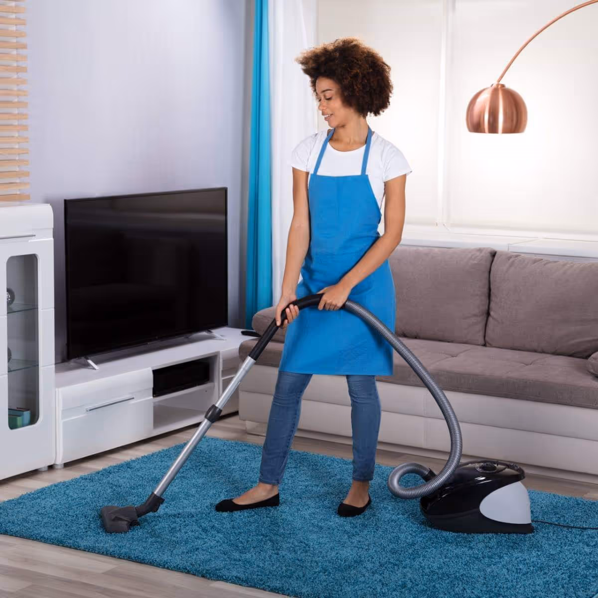 Caregiver in a blue apron vacuums a living room with sofa and TV.