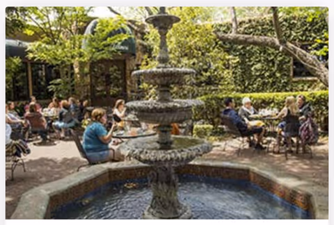 Tiered stone fountain in a shaded outdoor courtyard with people seated at tables around it.