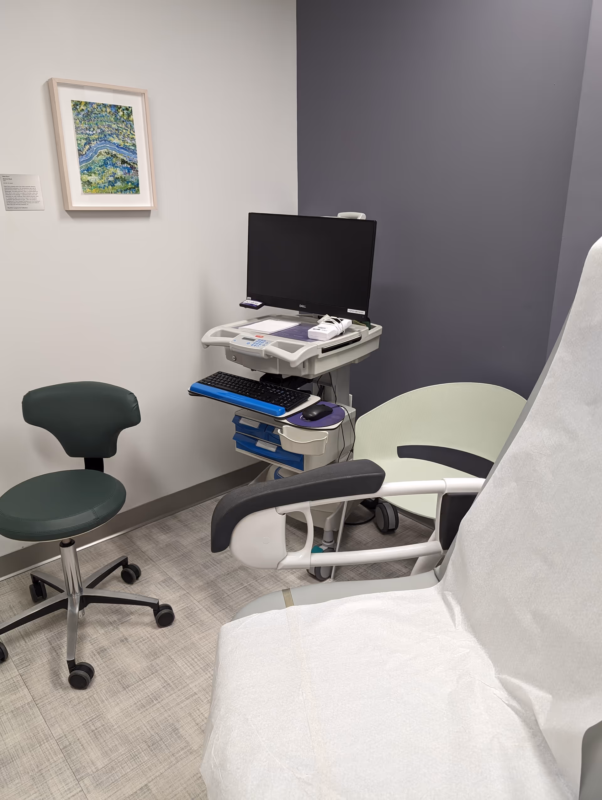 Healthcare examination chair with computer cart in a clinical room with neutral walls.