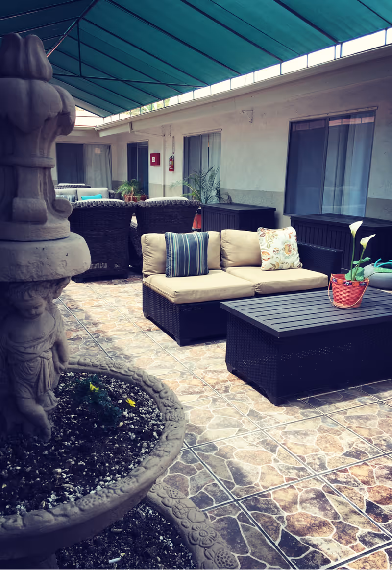 Covered outdoor patio area with cushioned wicker seating including a sofa with striped and floral pillows, a matching coffee table with a potted plant, and additional wicker chairs in the background. The floor is tiled with a stone pattern, and there is a decorative stone fountain planter with flowers in the foreground. The area is shaded by a green canopy and is adjacent to a building with windows and sliding doors.
