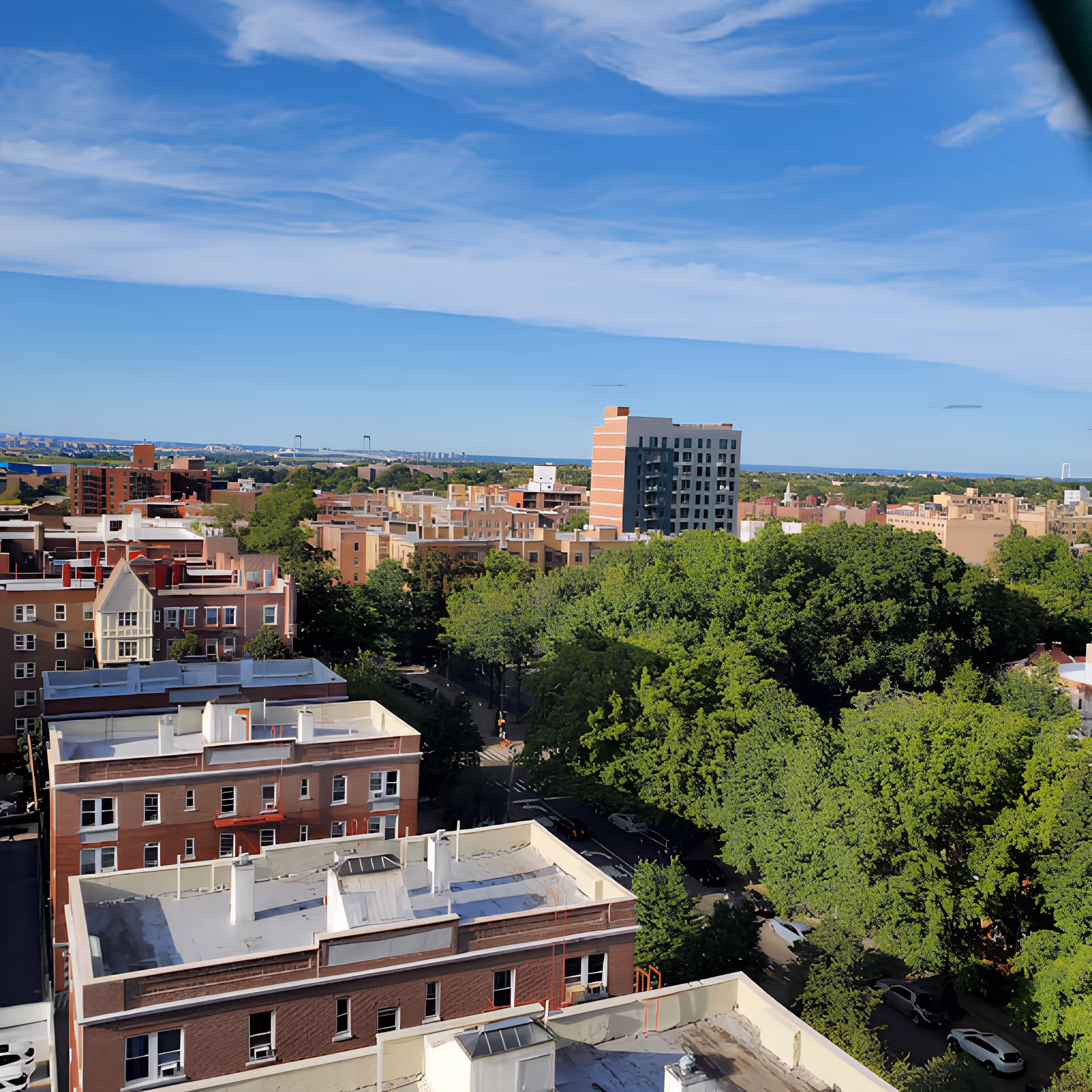 Aerial view of a cityscape featuring multiple residential buildings with flat rooftops, surrounded by dense green trees under a blue sky with scattered clouds.