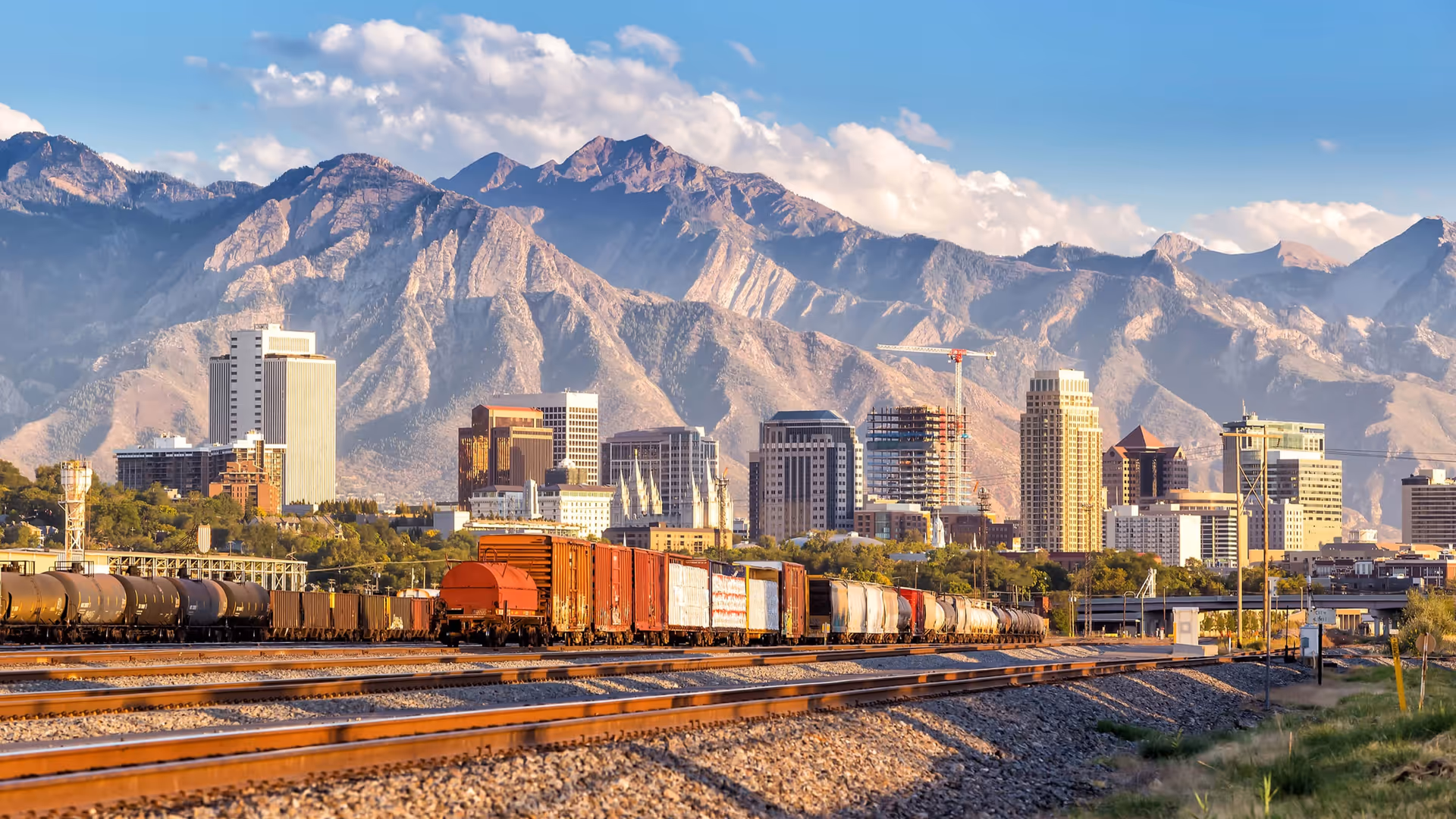 A city skyline with multiple tall buildings set against a backdrop of large, rugged mountains under a partly cloudy blue sky. In the foreground, there are several train tracks with freight train cars, including tankers and boxcars.