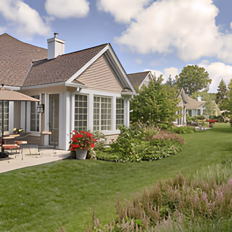 A landscaped lawn and patio beside a row of single-story residential units with large windows under a partly cloudy sky.