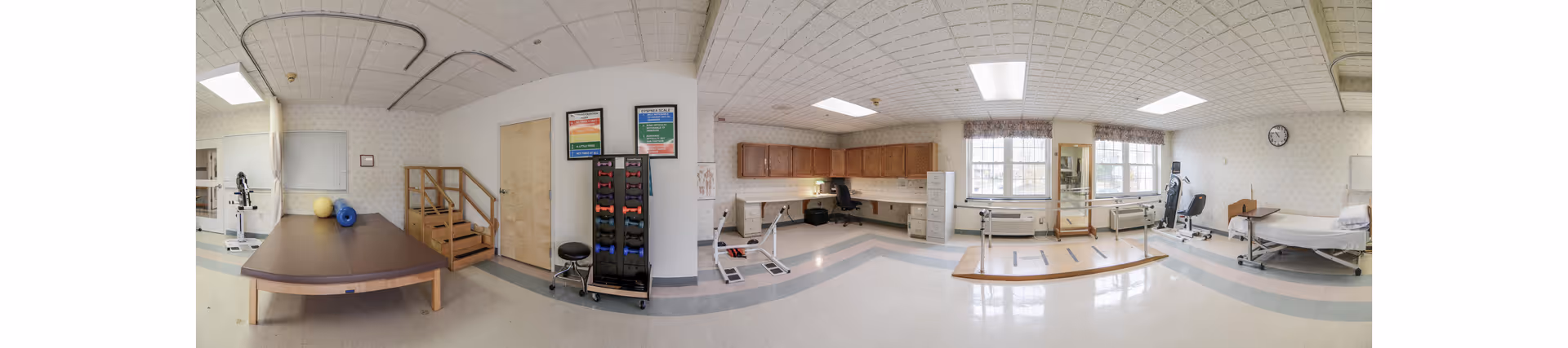 Wide interior of ManorCare Yeadon rehab area with therapy equipment, tables, and hospital beds visible.