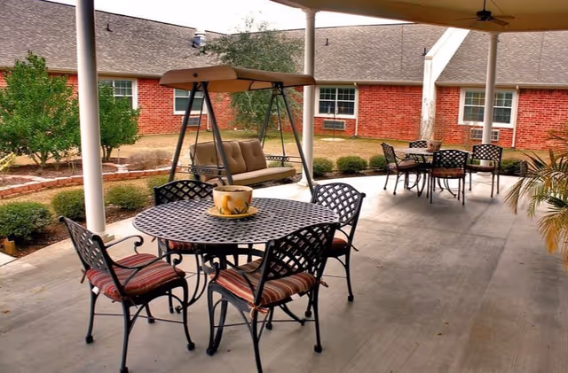 Covered outdoor patio area with metal tables and chairs with striped cushions, a cushioned swing, and potted plants. The patio overlooks a garden with bushes and a red brick building in the background.