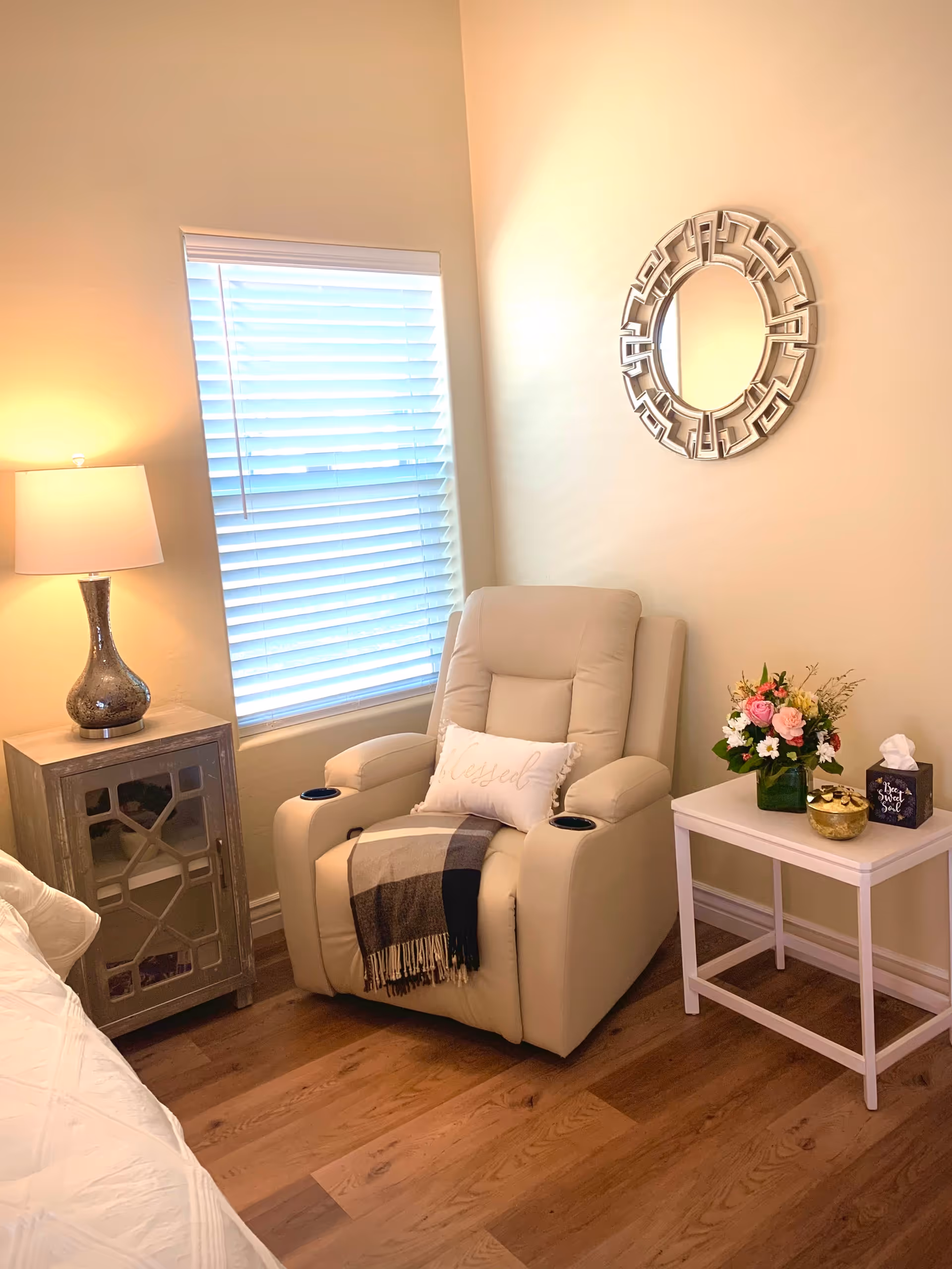 A cozy corner of a bedroom featuring a beige recliner chair with a 'blessed' pillow and a plaid throw blanket. To the left of the chair is a small wooden nightstand with a decorative lamp. To the right is a white side table holding a vase of colorful flowers, a gold decorative bowl, and a tissue box. A round decorative mirror hangs on the wall above the chair, and a window with white blinds is visible behind the chair.