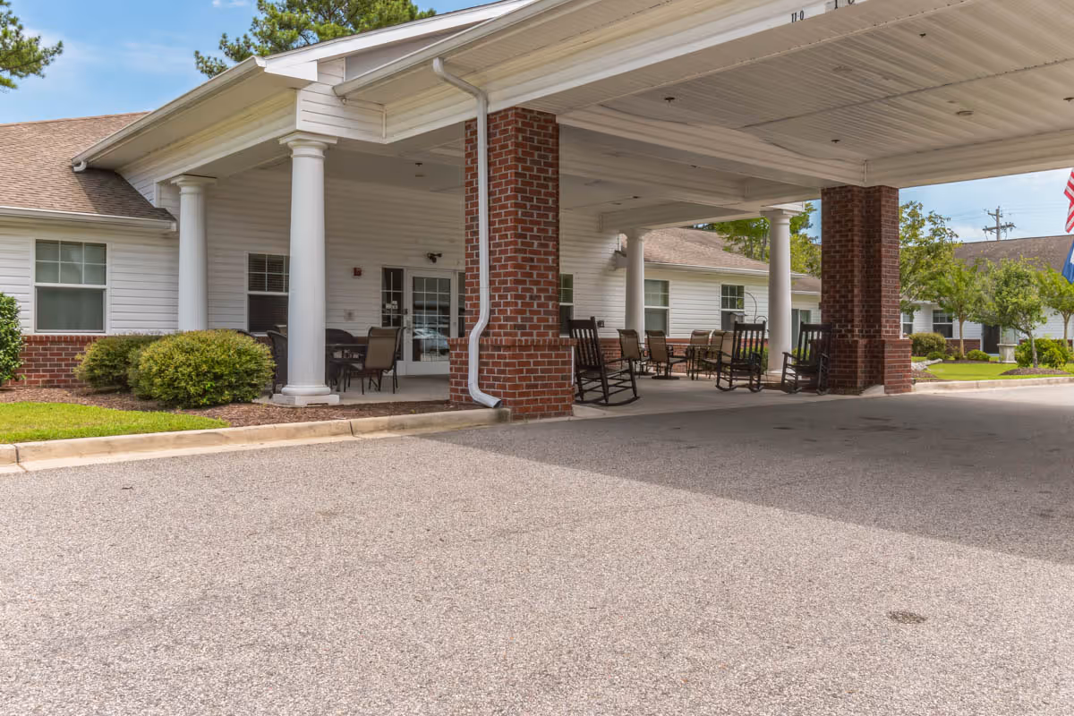 Covered entrance area of a senior living facility with white columns and brick pillars. Several rocking chairs and tables with chairs are arranged under the covered area. The building has white siding and multiple windows, with greenery and bushes around the perimeter.