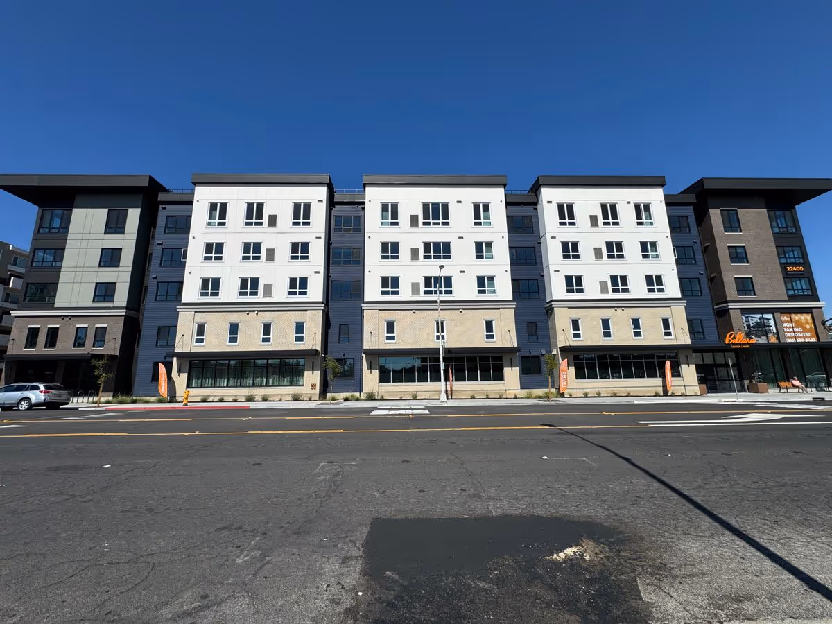 Front exterior of a multi-story senior living building with ground-floor storefronts along a wide street under a clear blue sky.