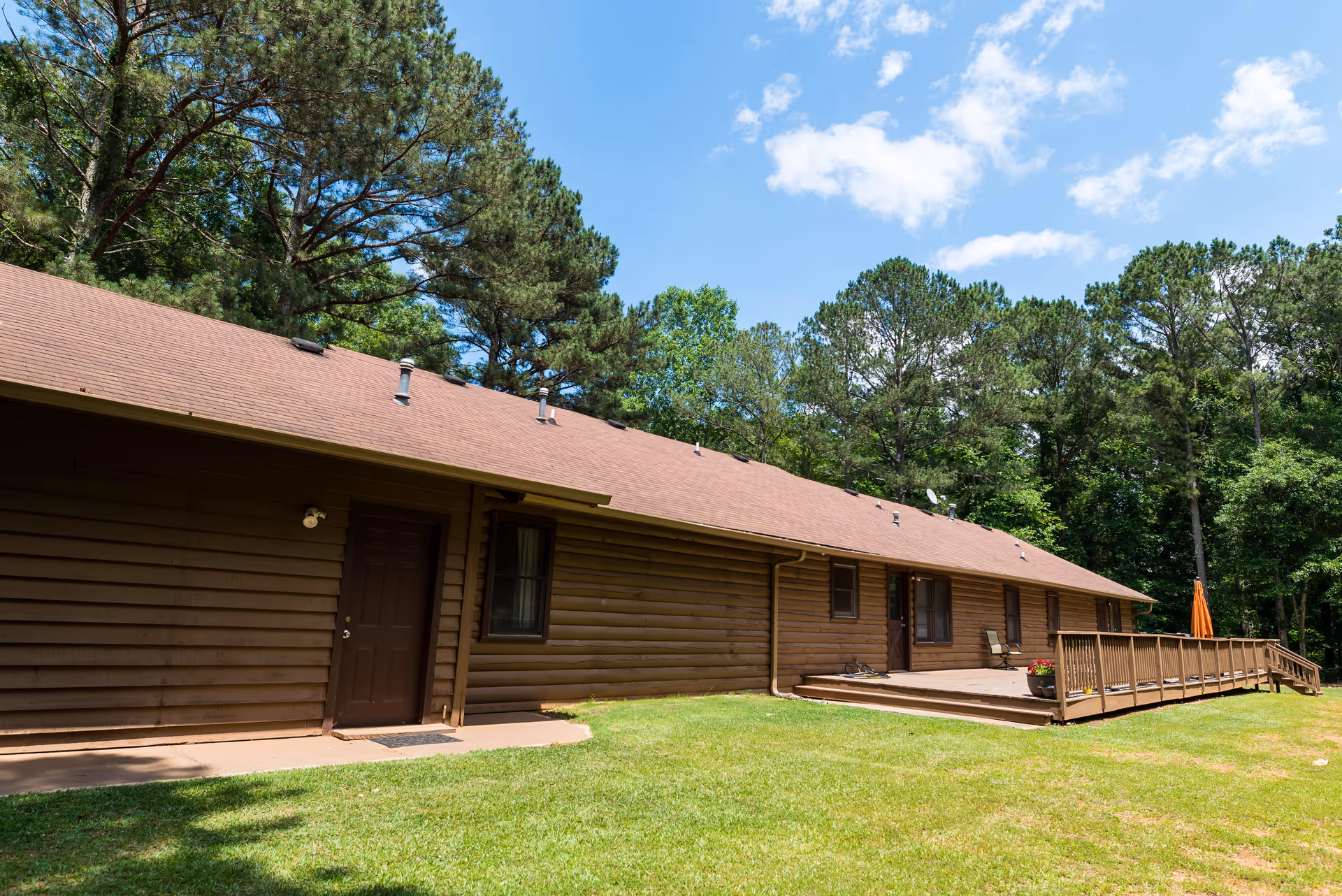 Exterior view of a single-story brown wooden building with a sloped roof, surrounded by green grass and tall trees under a blue sky with some clouds. The building has several doors and windows, and a wooden deck with a railing and an orange umbrella on the right side.