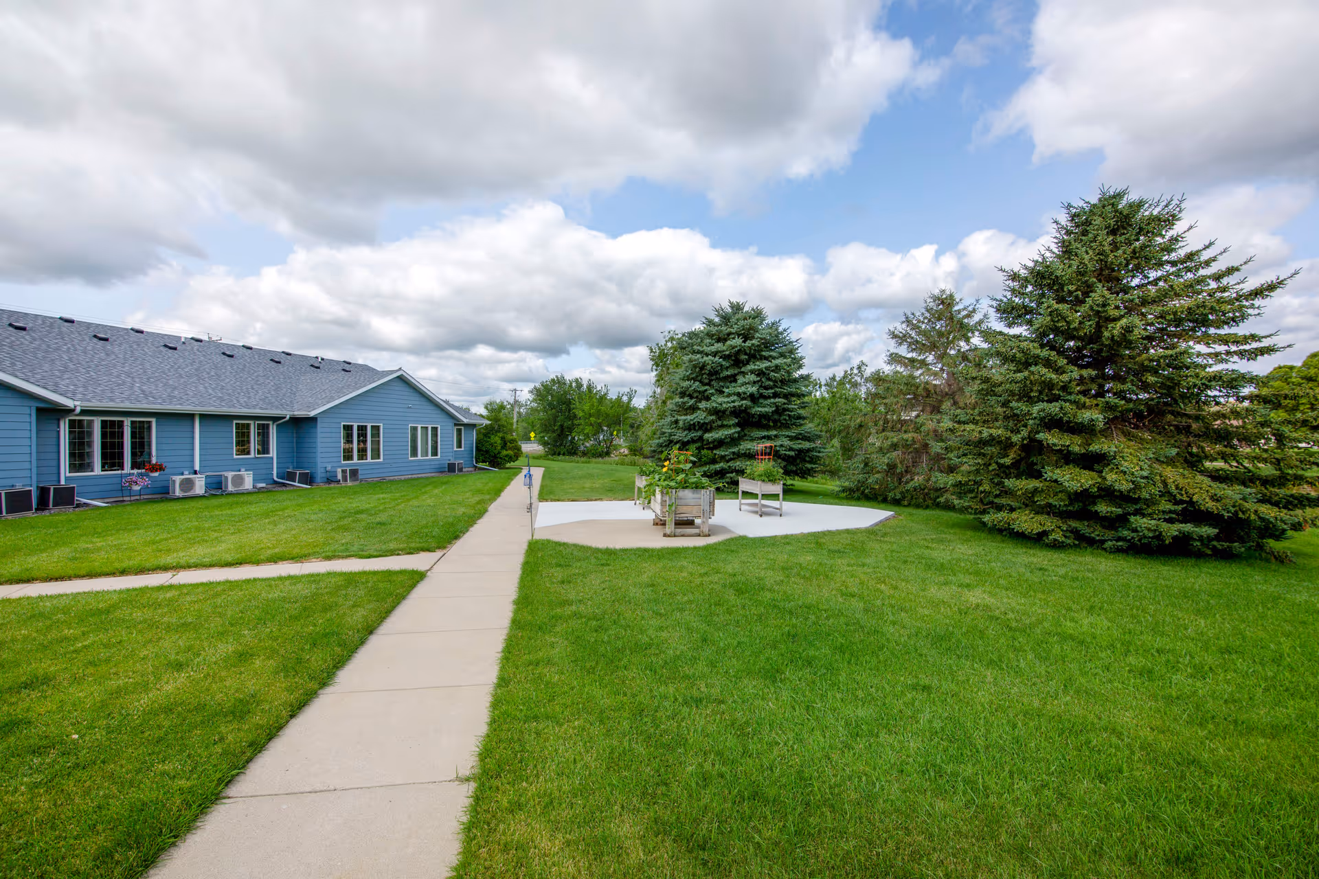 Blue senior living building with expansive green lawn, walkway, and circular outdoor seating area.