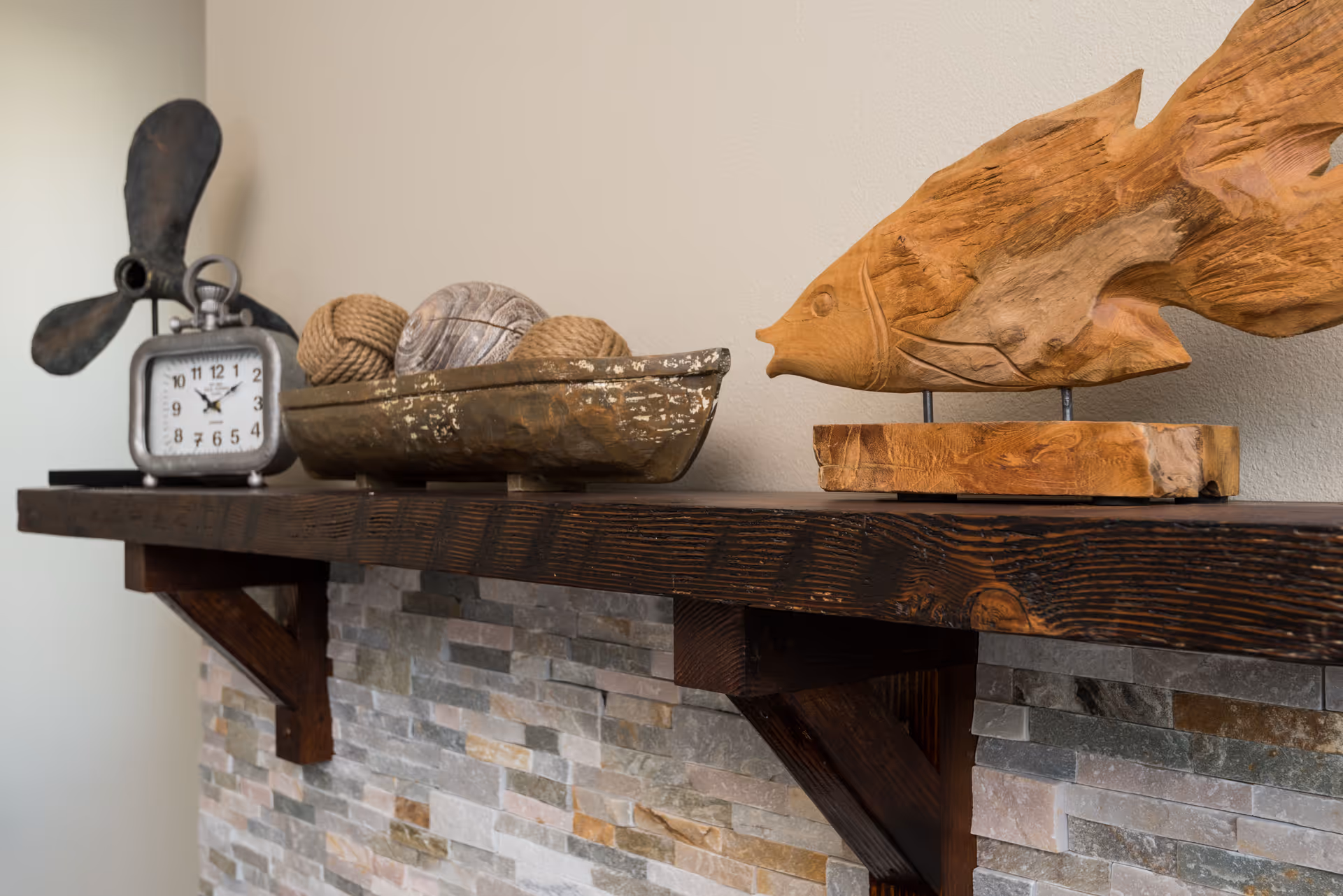 A close-up view of a wooden mantel shelf mounted on a stone wall. On the shelf, there is a decorative wooden fish sculpture, a rustic wooden bowl holding decorative balls, and a vintage-style clock with a propeller design behind it.