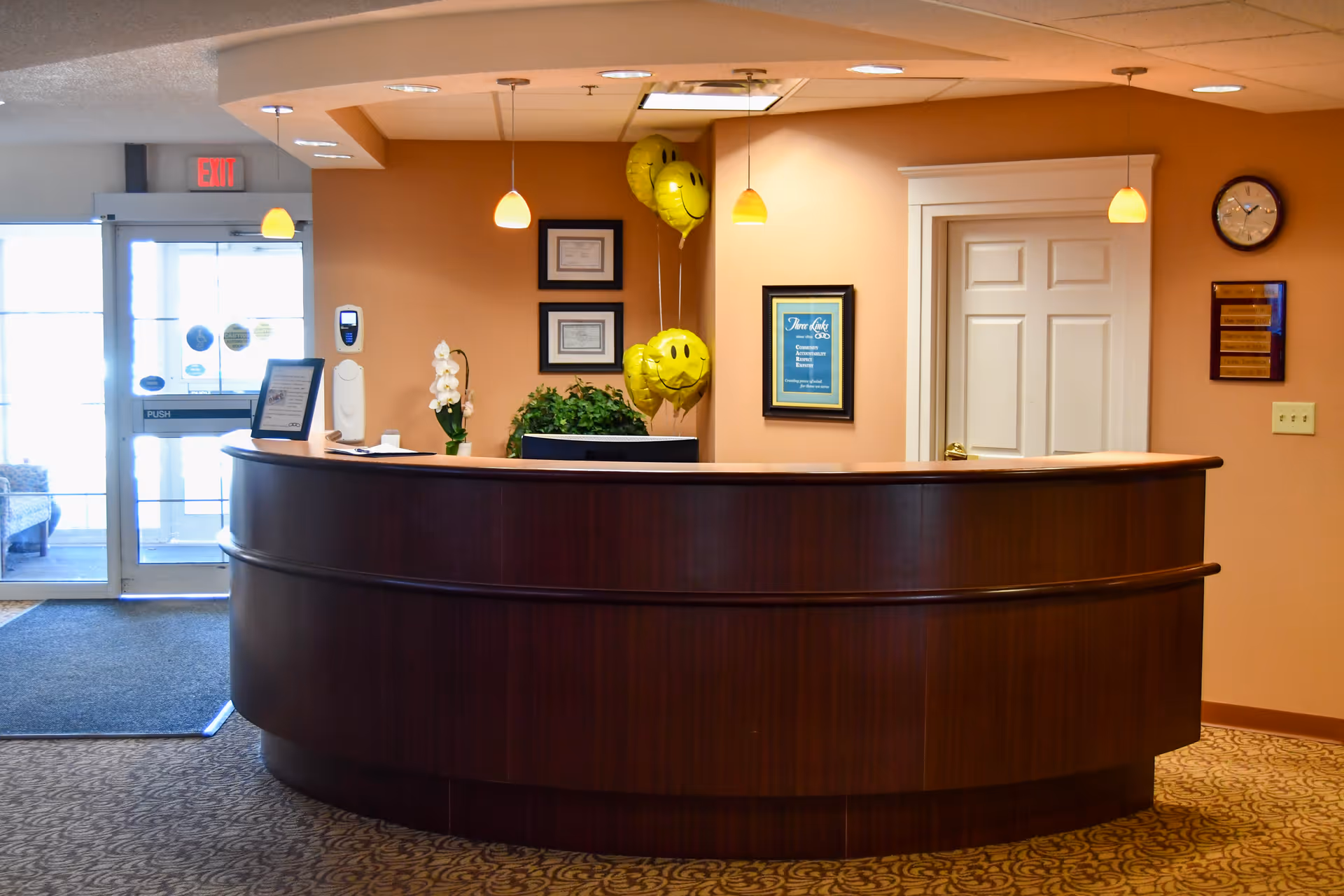 Reception area with a curved wooden desk, three yellow smiley face balloons, framed certificates and a clock on the wall, and a glass door entrance with an exit sign above it.