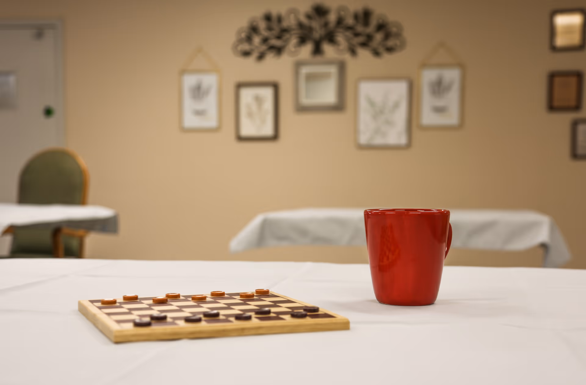 A table with a checkers board and a red mug in a communal dining/activity room with framed wall art in the background.