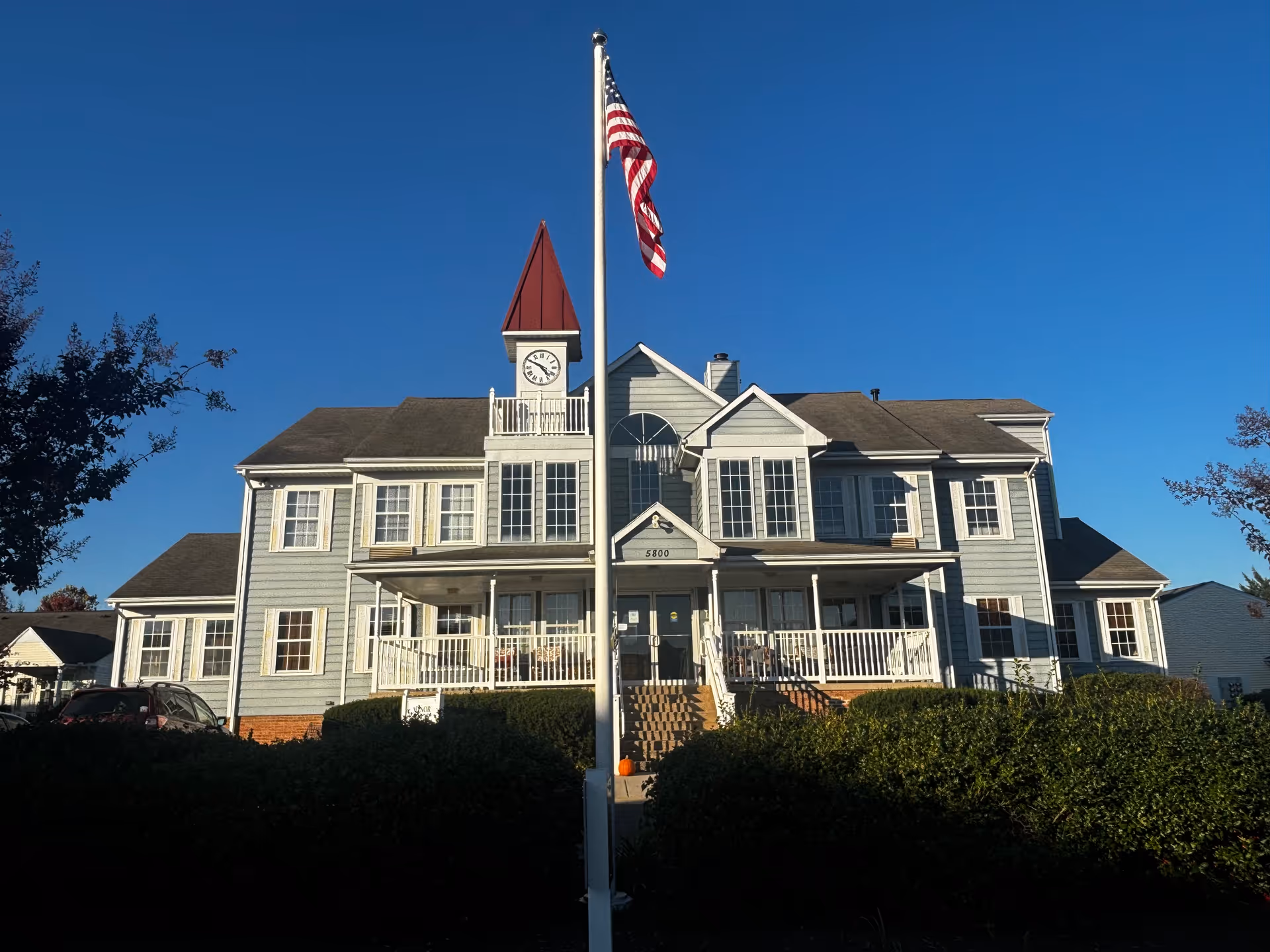 Front exterior of a two-story light-gray retirement community building with a central flagpole flying an American flag against a clear blue sky.