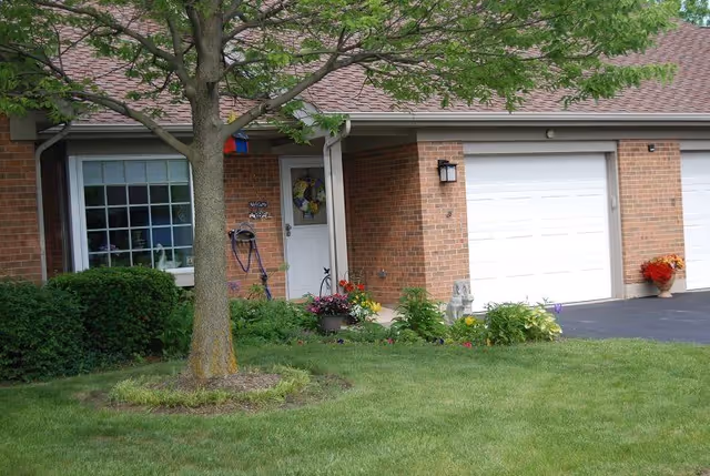 Brick single-story residential unit with a garage, front door, potted flowers and a tree on a green lawn.