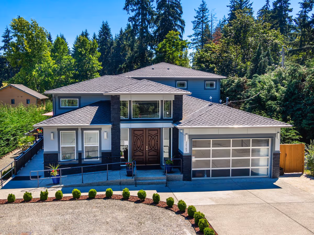 Two-story modern house with a double garage, central double doors, ramped entry, and landscaped front yard surrounded by trees.