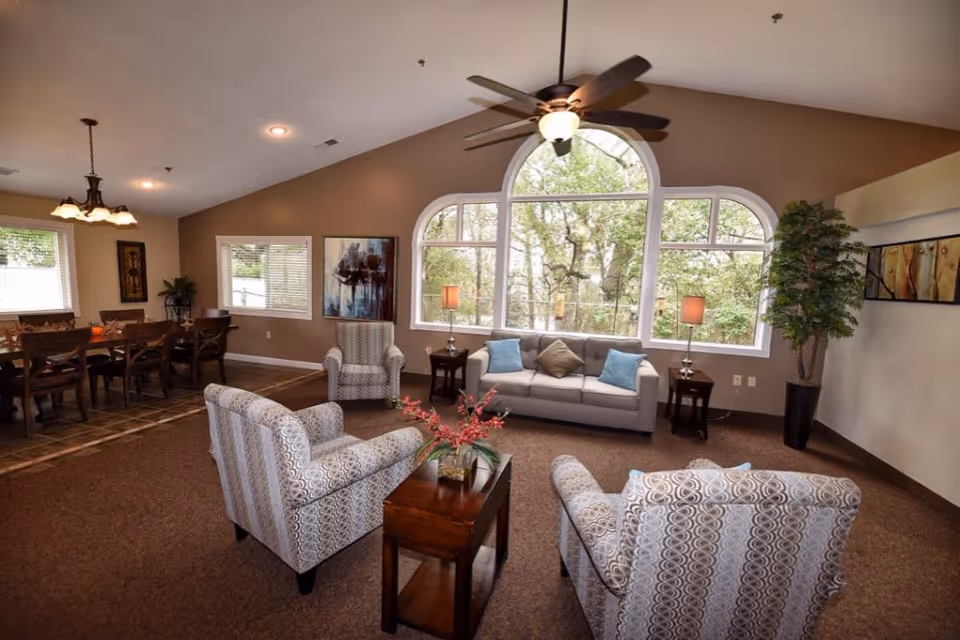 Spacious senior living lounge with sofas, armchairs, tables, a dining area, and a large arched window.