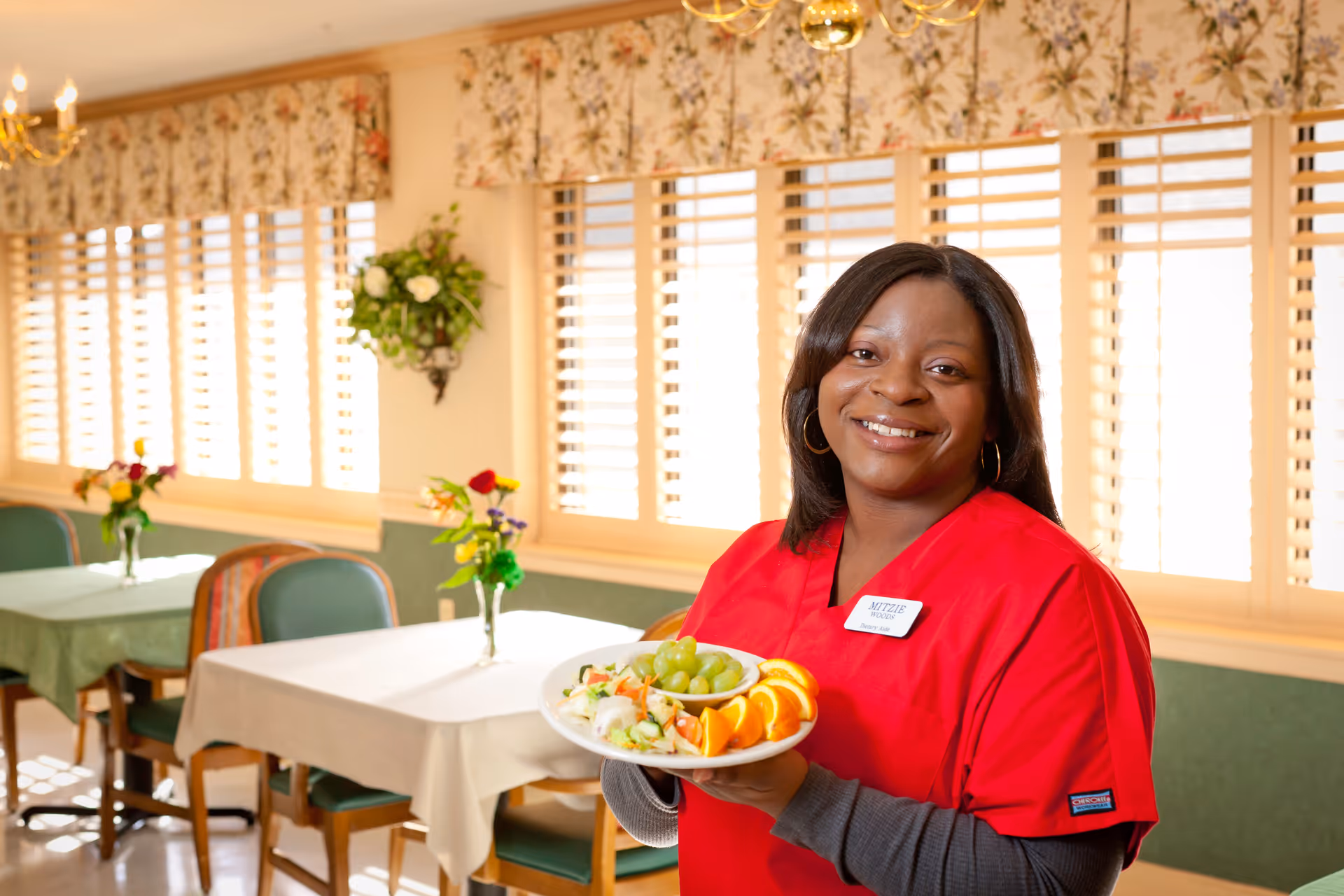 A smiling staff member in red scrubs holds a plate of fruit in a bright dining room with tables and floral decor.