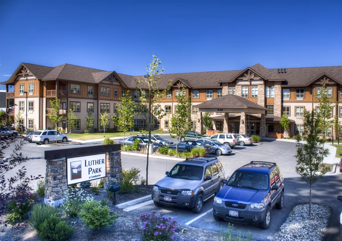 Exterior view of Luther Park at Sandpoint, a three-story senior living facility with a stone and wood facade. The building has multiple windows and a covered entrance. Several cars are parked in the parking lot in front of the building, and there are small trees and landscaping around the area under a clear blue sky.