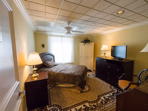 Cozy furnished bedroom with a bed, nightstands and lamps, a dresser with TV, armoire, ceiling fan, and patterned rug.