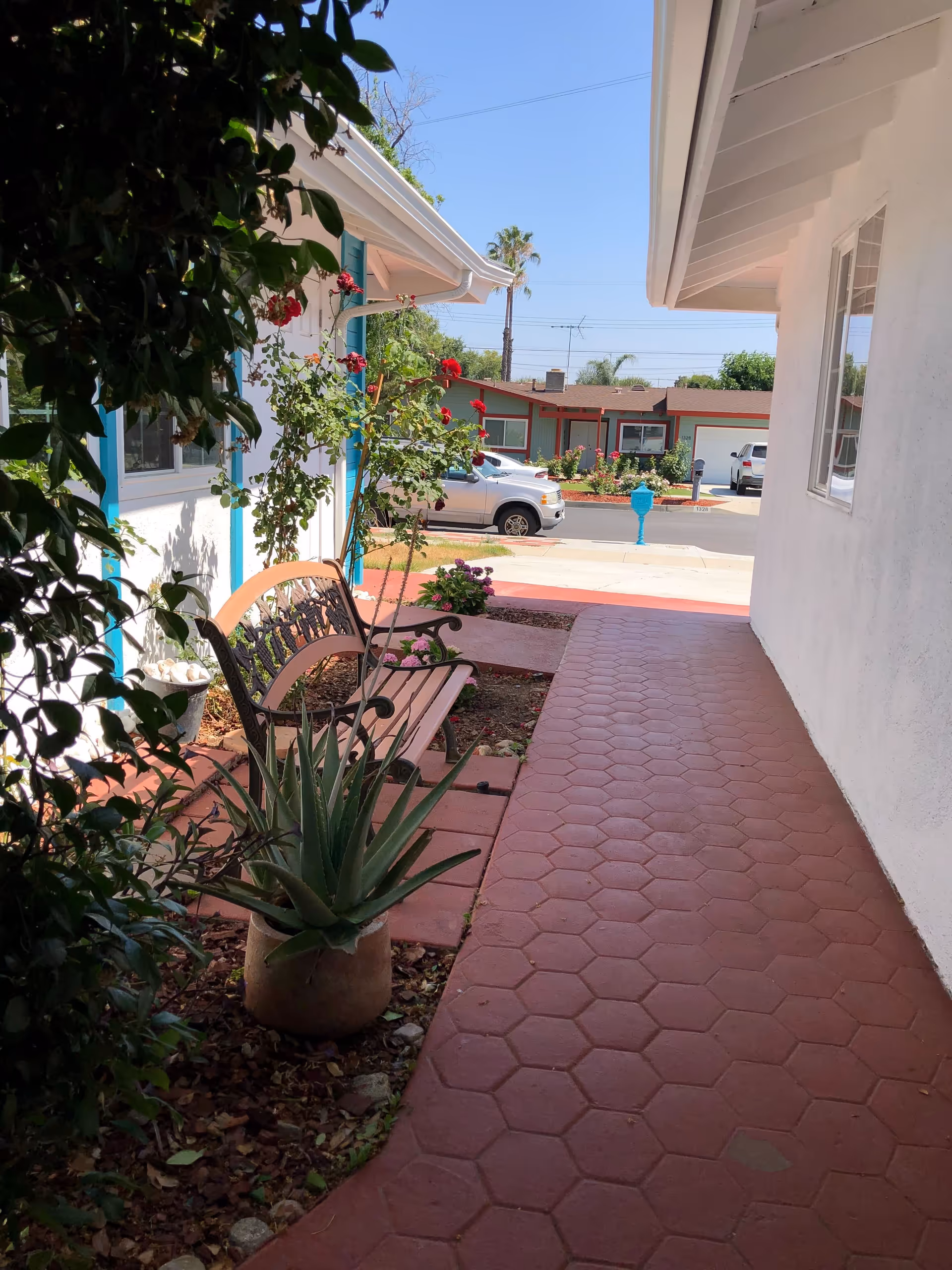 Shaded red-tiled walkway beside a white building with a bench, potted plants and flowers, opening to a street with parked cars and houses.