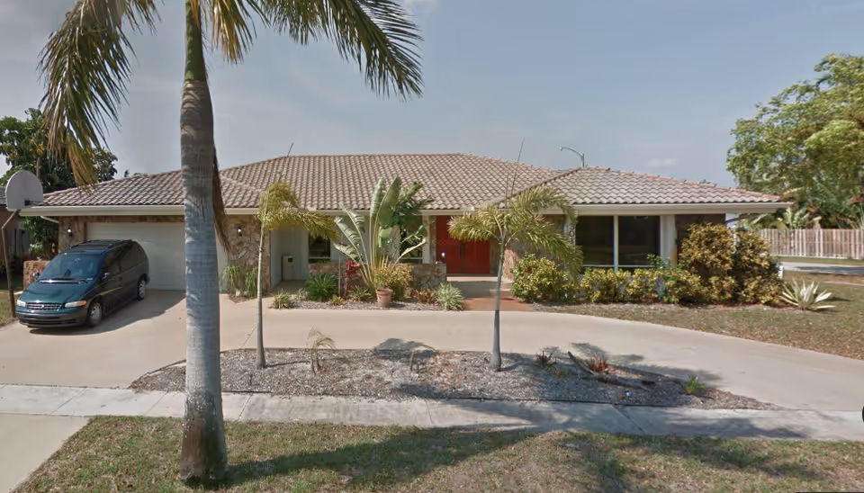 A single-story tan house with a circular driveway, palm trees, and a parked van.