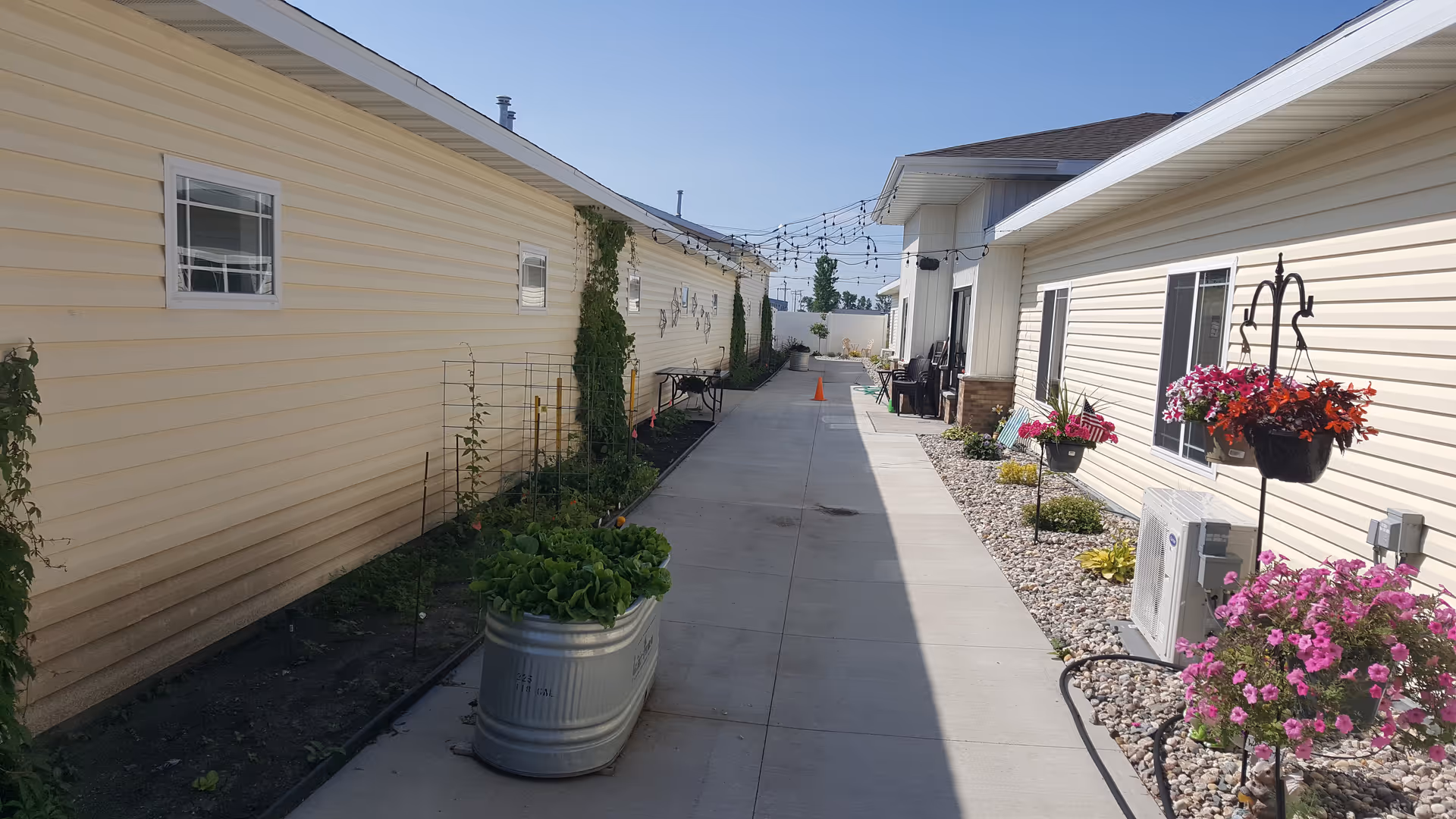Outdoor walkway between beige senior living buildings with string lights, planters, and seating at far end.