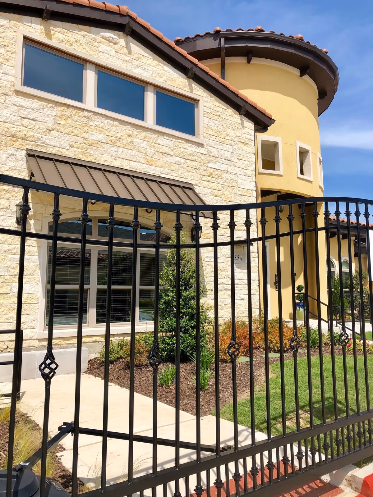 Stone building facade with curved tower, black wrought-iron fence, and landscaped grounds.