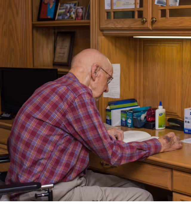 An elderly man wearing a red plaid shirt and glasses is sitting at a wooden desk reading a book. The desk has various office supplies including tape, glue, and boxes of paper. Behind the desk are wooden cabinets and shelves with framed photos and plaques.