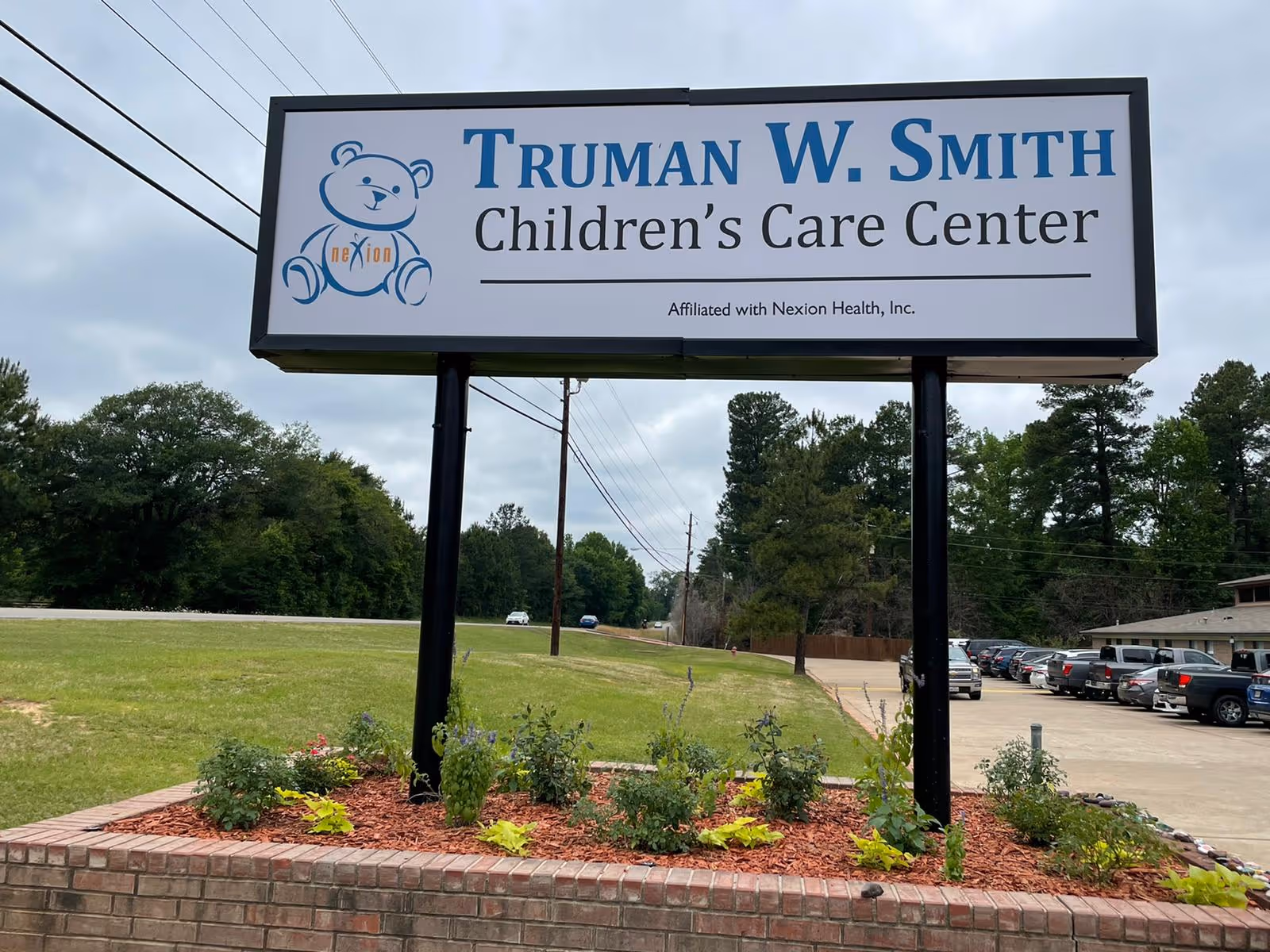 Outdoor view of a large sign for Truman W. Smith Children's Care Center with a teddy bear logo and affiliation with Nexion Health, Inc. The sign is mounted on black poles above a landscaped area with small plants and mulch. In the background, there is a parking lot with several vehicles and trees lining the horizon under a cloudy sky.