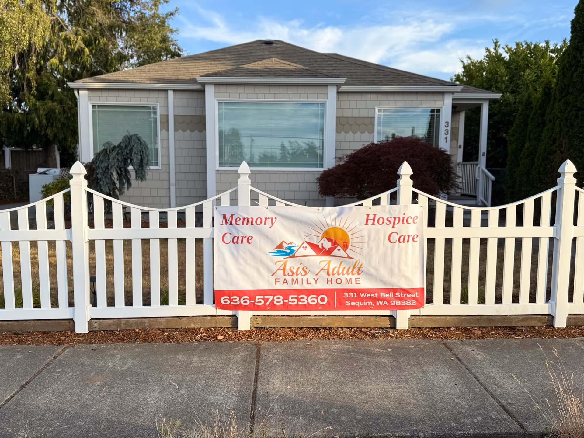 Front view of a small gray house with white picket fence and banner advertising memory care.
