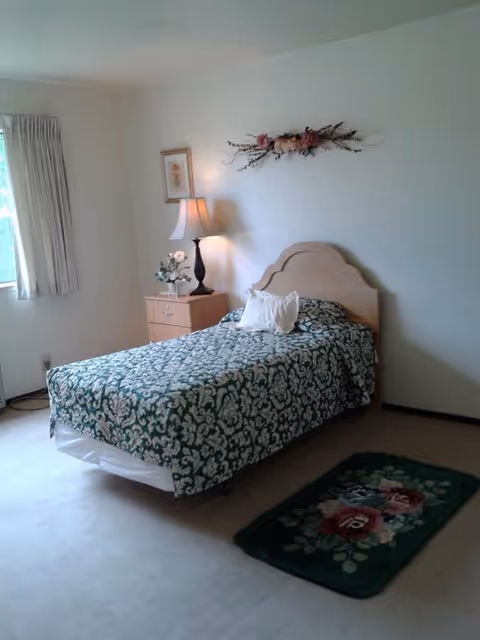 Single bed with green patterned bedspread, nightstand lamp, flowers, and floral rug in a small bedroom.