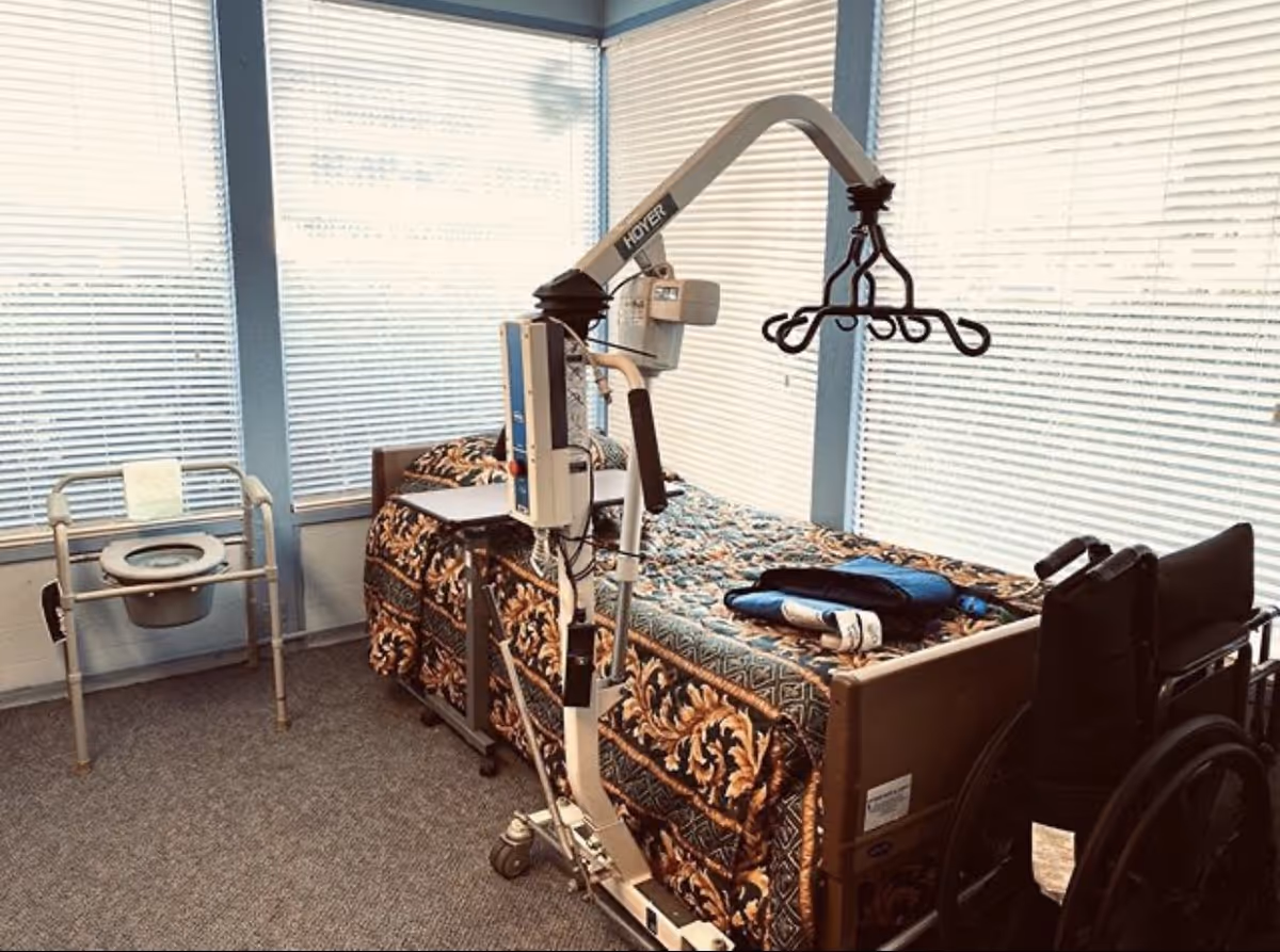 Hospital-style bed with lift device and wheelchair in a care room.