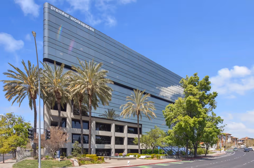 Modern glass office building with palm trees beside a curved road and blue sky.