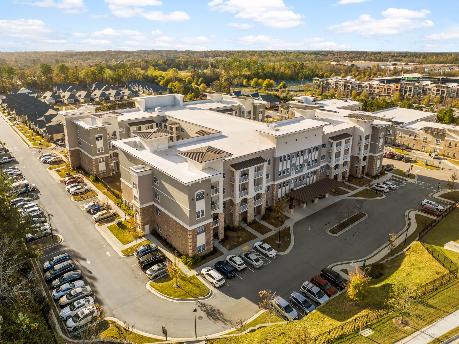 Aerial view of a large, modern senior living facility building with multiple floors and a spacious parking lot filled with cars. Surrounding the building are trees, residential houses, and other buildings under a partly cloudy sky.