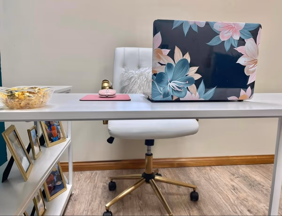 A white desk features a floral laptop, pink mouse pad, snack bowl, and photo frames.